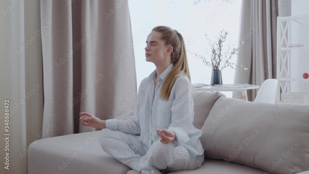 Peaceful young woman sitting in lotus pose on sofa in living room, meditating, attractive girl with closed eyes practicing yoga at home, stress relief concept, healthy good habit Peaceful young woman sitting in lotus pose on sofa in living room, meditating, attractive girl with closed eyes practicing yoga at home, stress relief concept, healthy good habit
