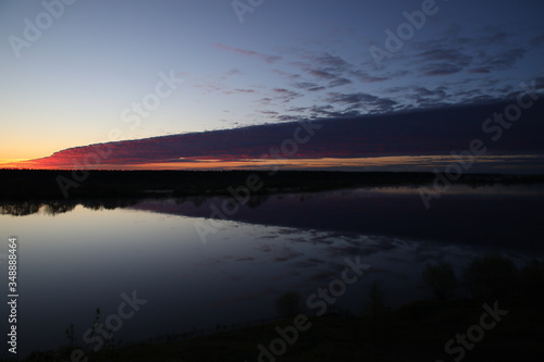 The water reflects the lilac sky the black forest by the river dividing the silvery surface from the clear horizon half covered with a mottled purple cloud in the mist at dawn.Landscape.Russia