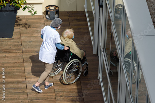 PARIS, FRANCE - MAY 13, 2020: nurse caring for an elderly and disabled woman in a nursing home on a sunny day during the coronavirus pandemic Covid-19. Both wear a protective face mask.