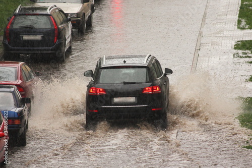 A dark SUV car drives fast with splash water on big puddle down the street next to parked vehicles in heavy rain in the city on a spring day  urban off-road back view