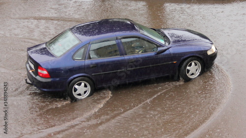 Dark blue sedan car rides carefully through a large puddle, heavy rain in the city