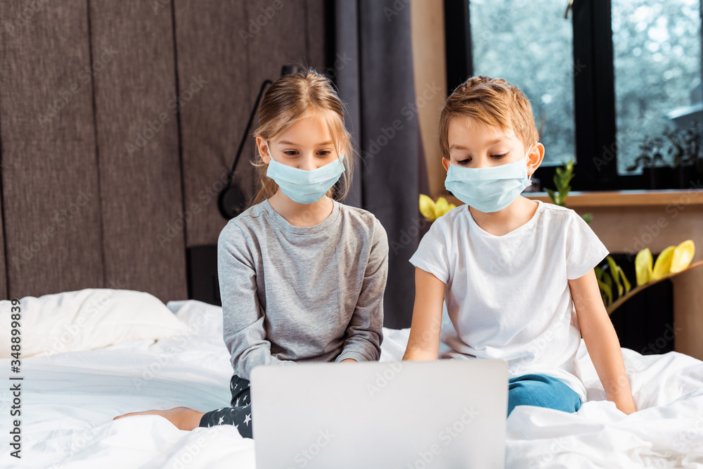 selective focus of kids in medical masks looking at laptop while e-learning in bedroom selective focus of kids in medical masks looking at laptop while e-learning in bedroom