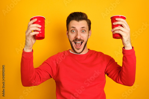 A hipster man with a beard opens his mouth, looks surprised, having fun on yellow background, holding red cups in both hands, wearing red sweatshot photo