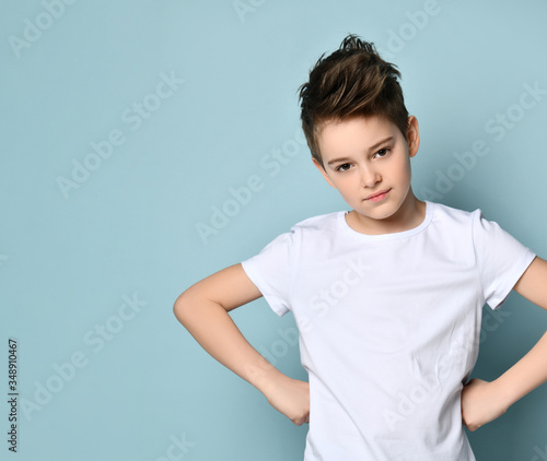 Scrappy brave teen boy in white t-shirt and jeans with stubborn look stands holding hands on hips. Close up portrait isolated on light blue