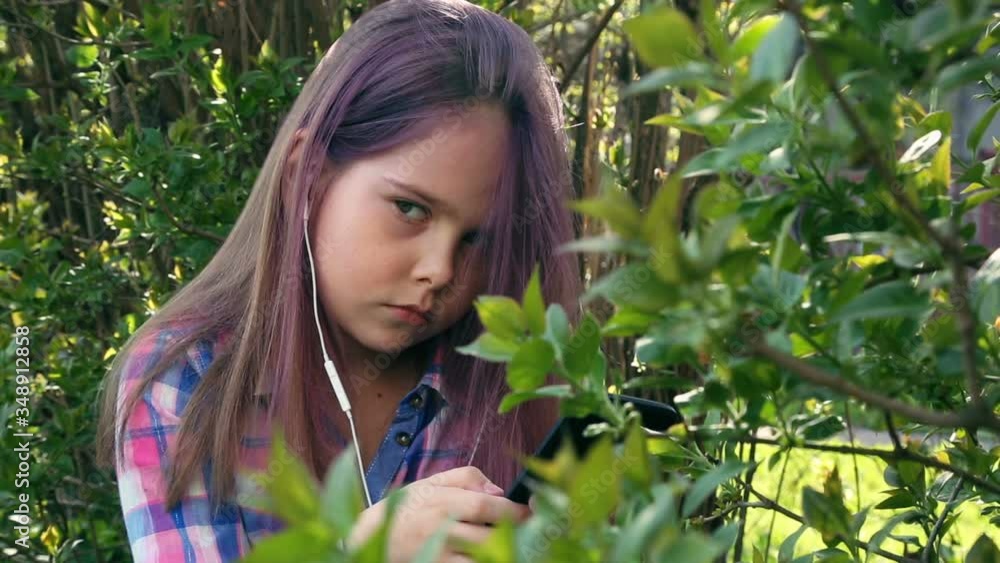 little beautiful girl plays on the phone and listens to music on the background of foliage shrubs and trees. little beautiful girl plays on the phone and listens to music on the background of foliage shrubs and trees.
