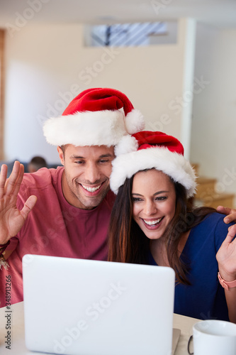 Hispanic Couple Wearing Santa Hats With Laptop Having Video Chat With Family At Christmas