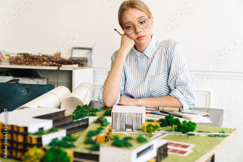 Photo of unhappy young woman architect designing draft with house model