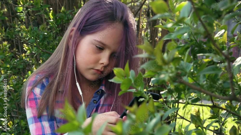 little beautiful girl plays on the phone and listens to music on the background of foliage shrubs and trees. little beautiful girl plays on the phone and listens to music on the background of foliage shrubs and trees.