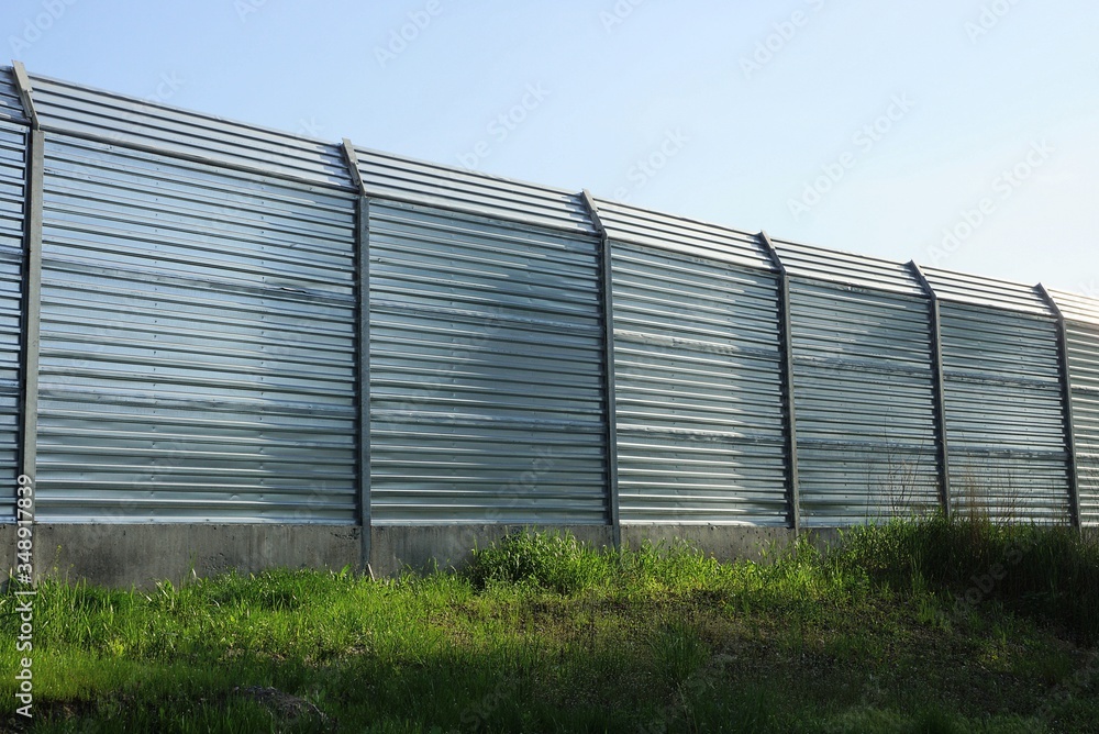 high gray metal fence wall in green grass on the street against a blue sky high gray metal fence wall in green grass on the street against a blue sky
