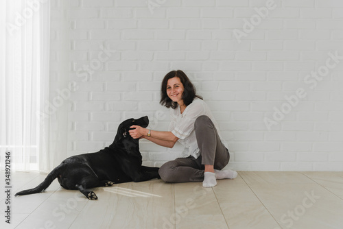 Portrait of a happy young adult in good mood sitting on a table at home with crossed legs. Happy housewife caresses a black labrador with pleasure. Copy space, family concept,middle-aged woman sits 
