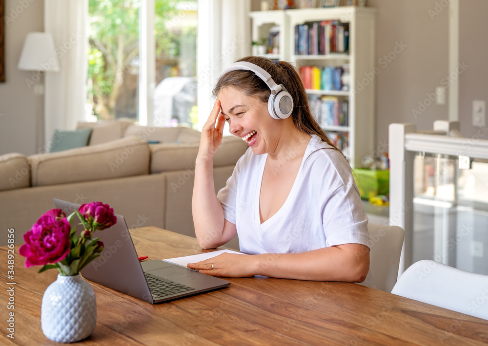 Happy positive girl with headphones working or studying online from home sitting at dining table laughing. Freelance, online work, learning and communication during quarantine concept. Home office. Happy positive girl with headphones working or studying online from home sitting at dining table laughing. Freelance, online work, learning and communication during quarantine concept. Home office.