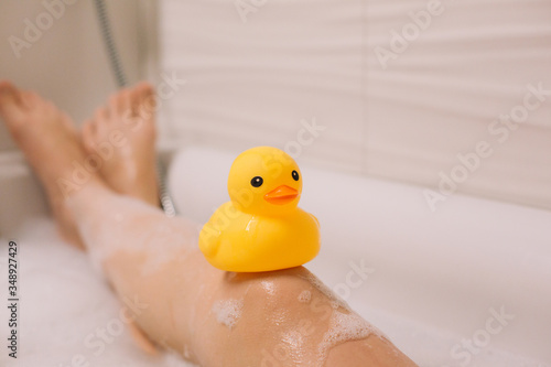 Girl is holding on her legs yellow rubber duck in the bath with bubble foam