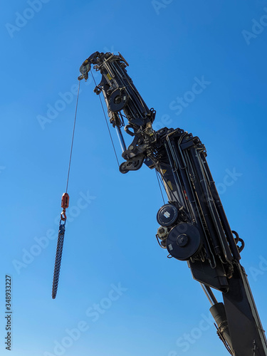 Closeup of extendable black truck crane with blue sky background. The photo was taken at a construction site and includes the hydraulic mechanism, the pipes, the crane hook and cable.
