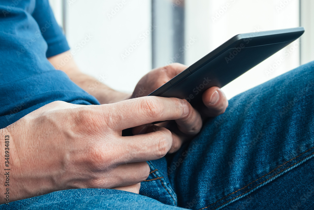 A man in a blue t-shirt and jeans holds an electronic book or tablet in his hands. Reading books at home by the window. Selective focus. Closeup view