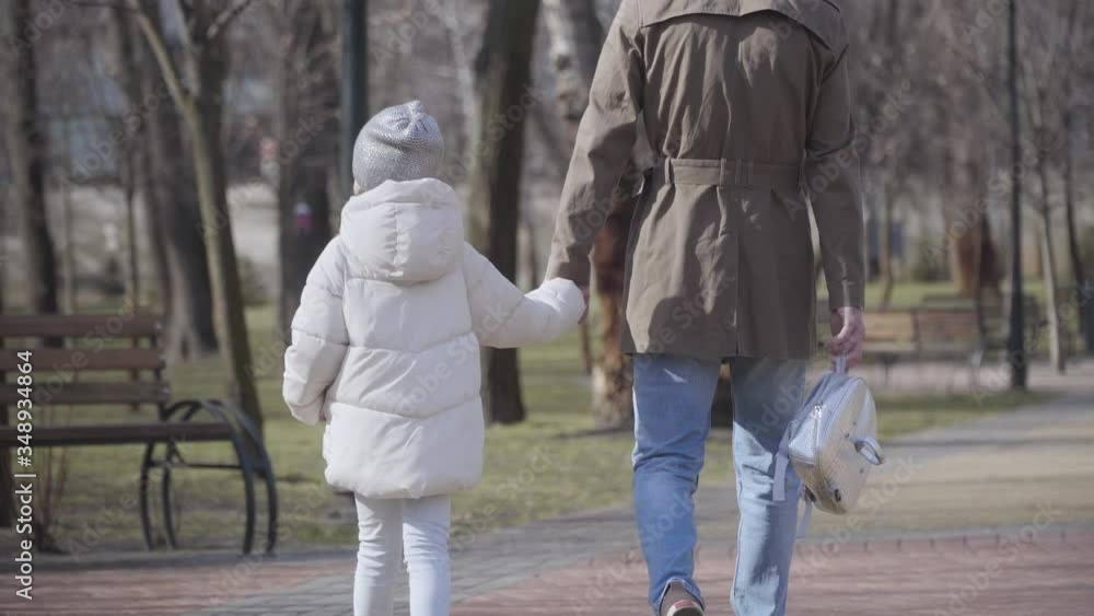 Back view of little schoolgirl walking with young father along alley in autumn or spring park. Girl in white coat strolling after studies with parent. Lifestyle, family, support. Back view of little schoolgirl walking with young father along alley in autumn or spring park. Girl in white coat strolling after studies with parent. Lifestyle, family, support.