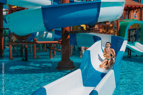 Obraz na płótnie Little girl in swimsuit goes down the blue slides to the pool, Mom and daughter play and swim in the outdoor pool