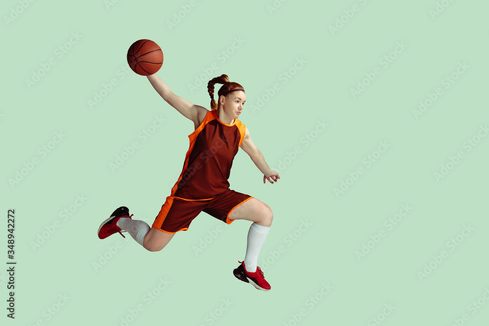 Young caucasian female basketball player in action, motion in high jump isolated on mint colored background. Concept of sport, movement, energy and dynamic, healthy lifestyle. Training, practicing.
