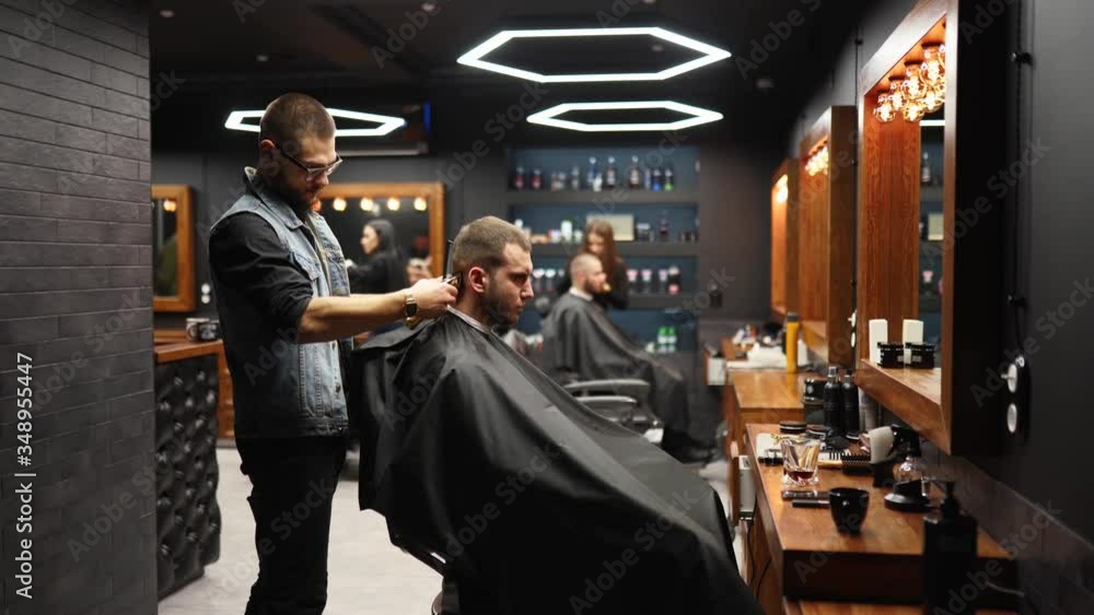 Trendy barber cuts bearded man's hair with a clipper in barbershop. Men's hairstyling and hair cutting in salon. Grooming the hair with trimmer. Hairdresser doing haircut in retro hair salon. Tracking Trendy barber cuts bearded man's hair with a clipper in barbershop. Men's hairstyling and hair cutting in salon. Grooming the hair with trimmer. Hairdresser doing haircut in retro hair salon. Tracking