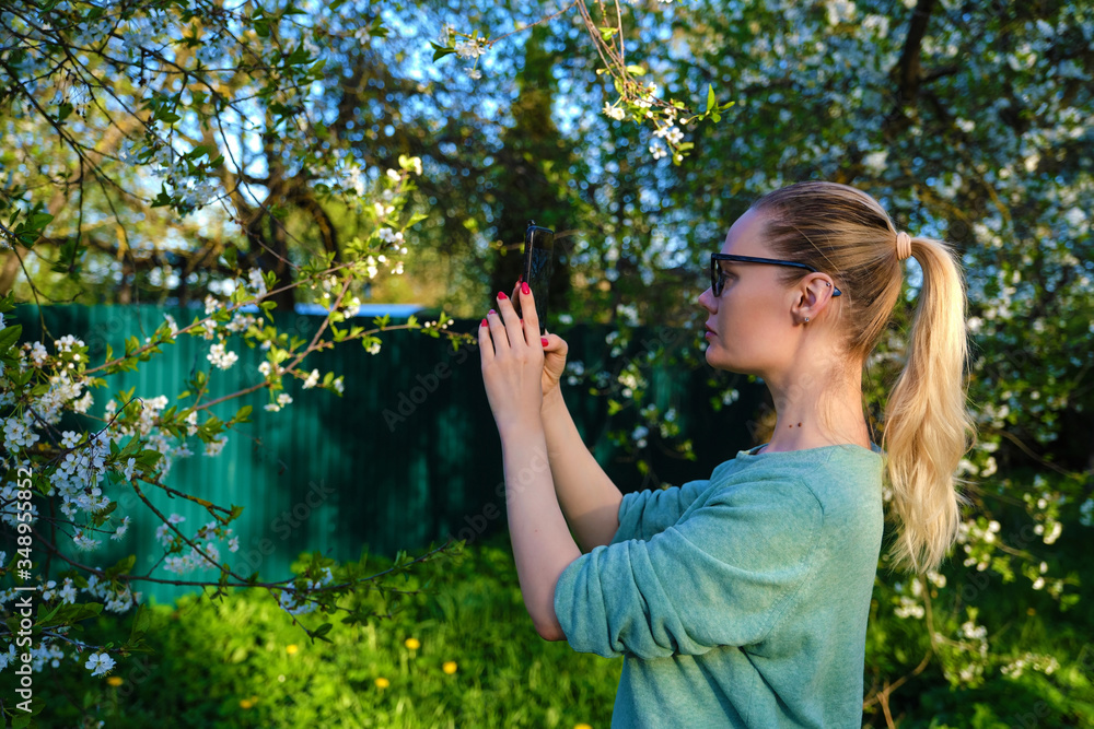 Blond girl in glasses is photographing flowering trees in May with her phone. Blond girl in glasses is photographing flowering trees in May with her phone.