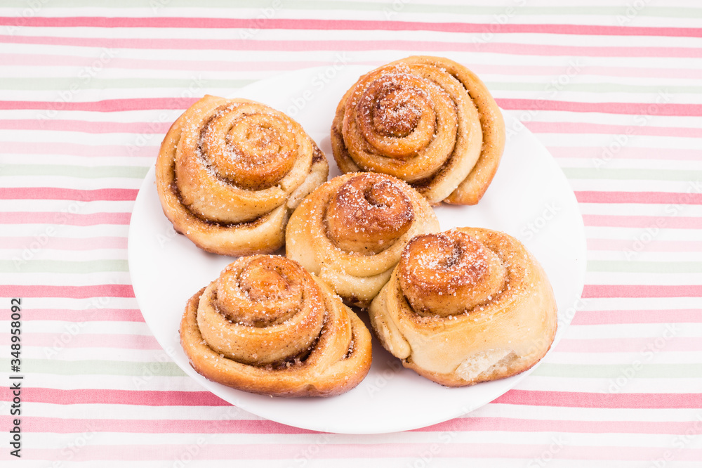 Freshly baked homemade snail buns with sugar and cinnamon on white plate and striped tablecloth. Balanced nutrition, proteins and carbohydrates, cereals