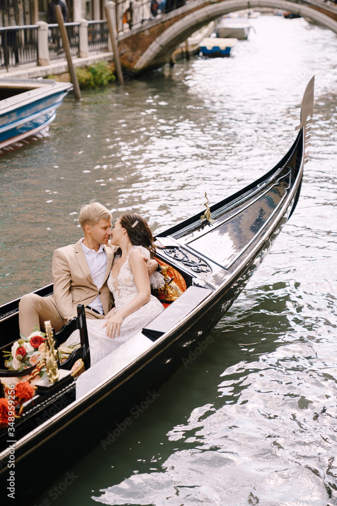 Italy wedding in Venice. The bride and groom ride in a classic wooden gondola along a narrow Venetian canal. Close-up of cuddles newlyweds.