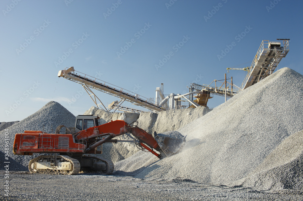 Powerful red excavator with a full bucket of crushed stone on a background of constructions of mining enterprise. Heavy mining equipment. Powerful red excavator with a full bucket of crushed stone on a background of constructions of mining enterprise. Heavy mining equipment.