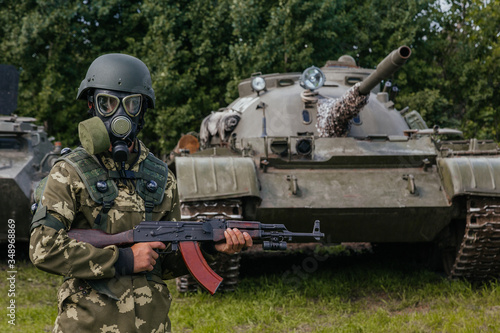 A soldier in a gas mask with a Kalashnikov rifle against the background of armored vehicles.