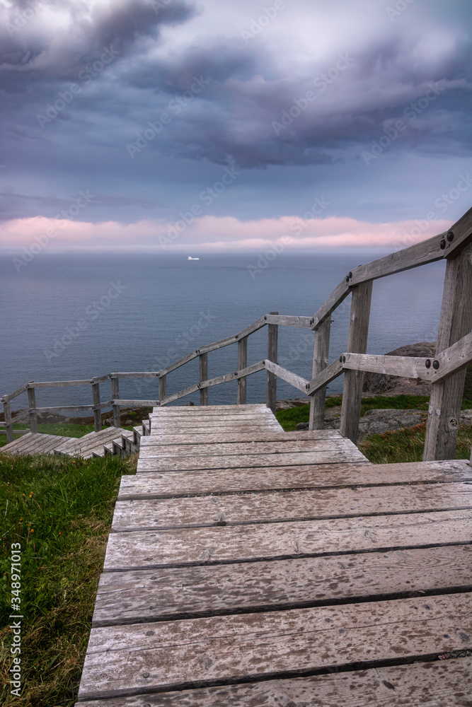 Naklejka premium A wooden staircase leading down to a dramatic coastal seascape with ominous clouds and fog rolling in. An iceberg in the ocean off shore. North Head Trail at Signal Hill - Newfoundland, Canada.