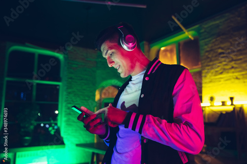 Music. Cinematic portrait of stylish man in neon lighted interior. Toned like cinema effects, bright neoned colors. Caucasian model using gadgets, devices in colorful lights indoors. Youth culture.