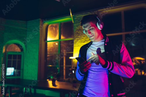 Choosing song. Cinematic portrait of stylish man in neon lighted interior. Toned like cinema effects, bright neoned colors. Caucasian model using gadgets in colorful lights indoors. Youth culture.
