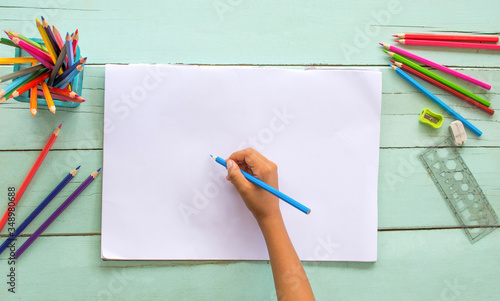 The top view of a child's hand, using colored pencils, drawn on white paper on a wooden table.