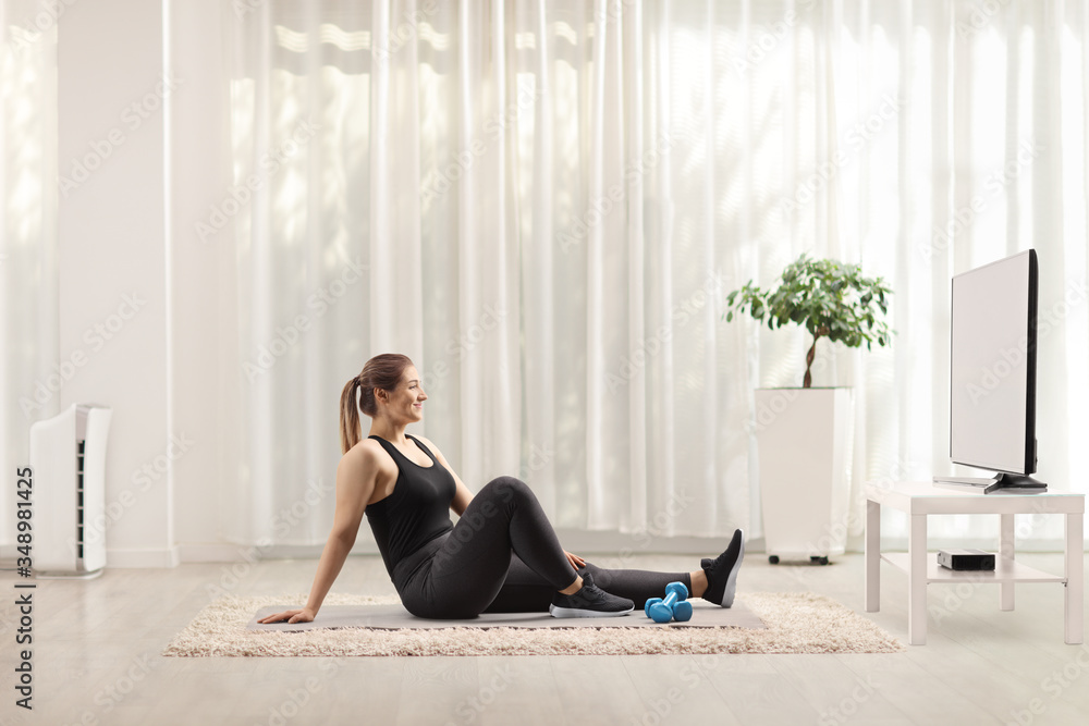 Young woman sitting on an exercise mat with dumbbells and watching tv Young woman sitting on an exercise mat with dumbbells and watching tv