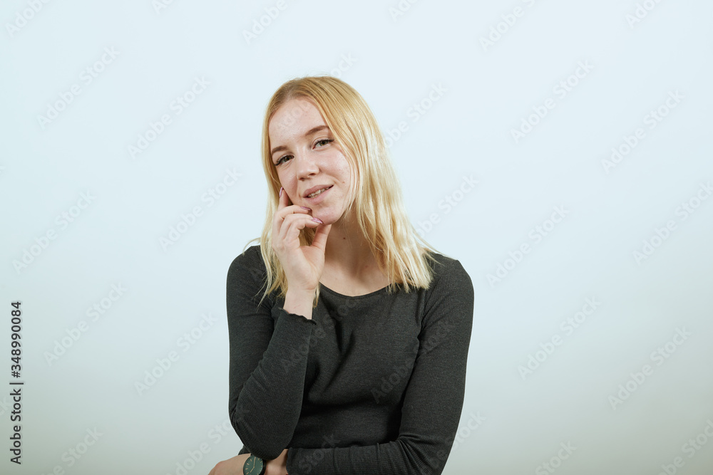 Young Blonde Woman In Black Sweater With Stylish Watch On White Background, Dreamy Girl Holds Her Hand To Chin And Thinks. The Concept Of Smart People. Young Blonde Woman In Black Sweater With Stylish Watch On White Background, Dreamy Girl Holds Her Hand To Chin And Thinks. The Concept Of Smart People.