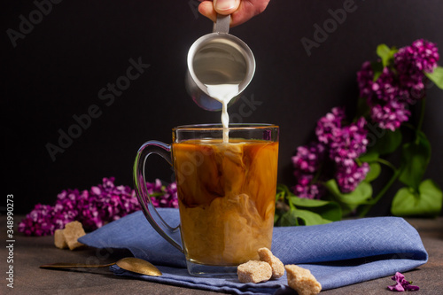 Cup with coffee with pouring cream, milk on a table with lilac flowers in the background