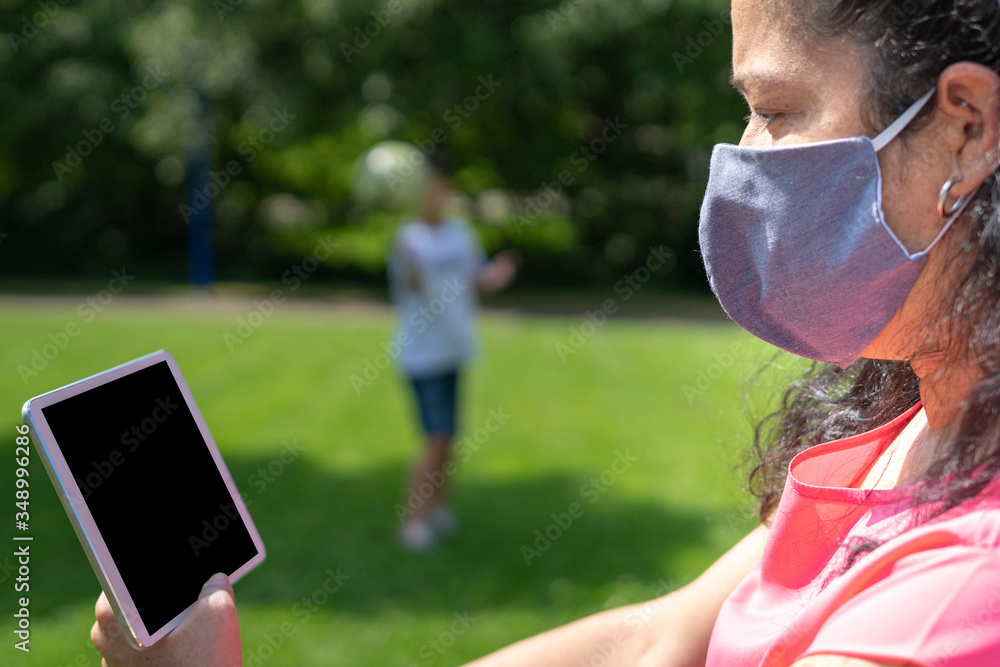 Mother reads her tablet very concentrated in the park with a mask covering her mouth while her child plays with the ball. Enjoy the outdoors. Mother reads her tablet very concentrated in the park with a mask covering her mouth while her child plays with the ball. Enjoy the outdoors.