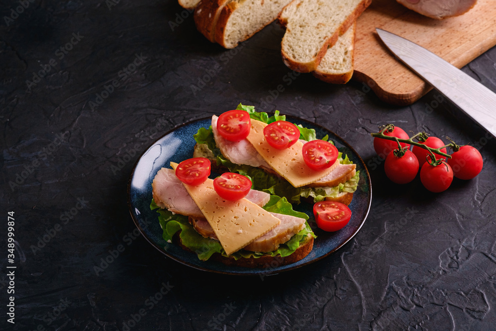 Sandwiches with turkey ham meat, green salad, cheese and fresh cherry tomatoes slices on blue plate near to ingredients on cutting board and kitchen knife, dark textured background, angle view