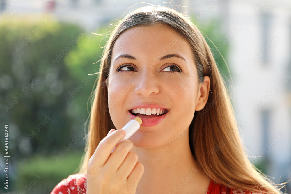 Fototapeta premium Portrait of a happy young woman applying lip balm in summer time outdoor