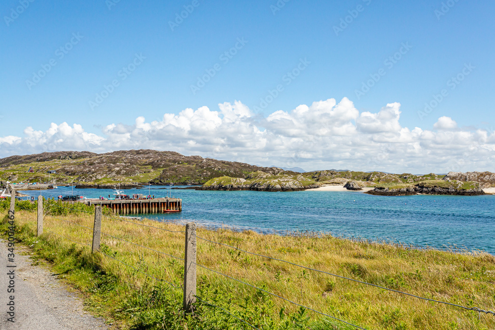 Seafront promenade with wire fence with bay, pier and rocky hills in background, sunny spring day on Inishbofin Island, County Galway, Ireland