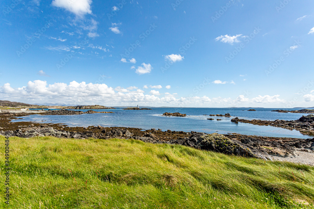 Shoreline with seaweed on the stones with the Atlantic Ocean in the background seen from a meadow with green grass, sunny spring day on Inishbofin Island, County Galway, Ireland