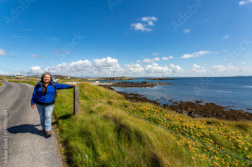 Mature Mexican woman in blue jacket on the coastal road with dramatic shoreline scenery, sunny spring day with blue sky and white clouds on Inishbofin Island, County Galway, Ireland photo