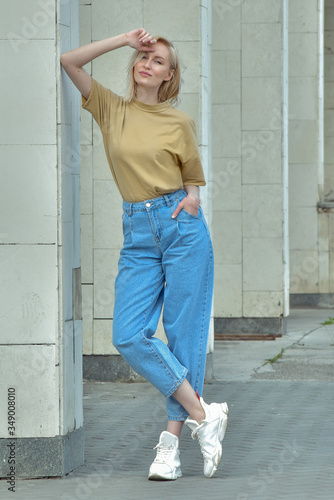 Young attractive beautiful fashionable slim woman is standing near a gray wall in bright blue jeans and a beige t-shirt. wind in the hair. modern woman in sneakers. street style