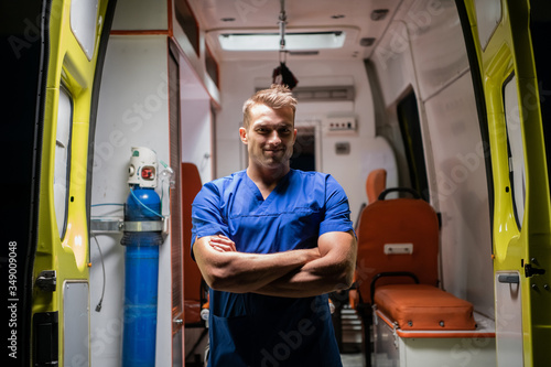 A handsome man in a medical uniform looking at the camera with a smile, arms folded, ambulance car in the background