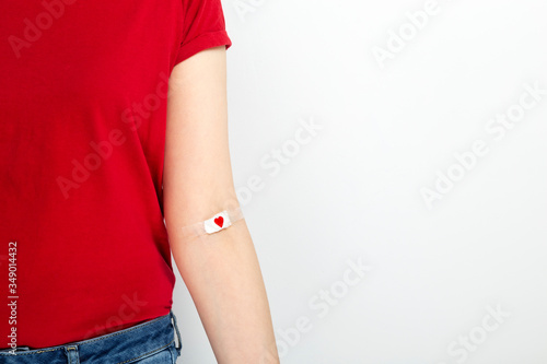 Blood donorship. Young girl in red T-shirt hand taped with patch with red heart after giving blood on gray background. Copy space photo