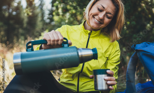 Girl smiles drink coffee from thermos on nature after training, woman hold hand mug of warm tea outdoors, hiker enjoys breakfast rest in green forest. Tourist relax while traveling photo