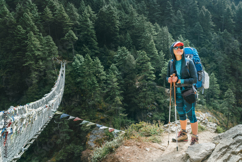 Young smiling female photographer walking Everest Base Camp route at canyon over Suspension Hillary Bridge with multicolored Tibetan Prayer flags hinged over gorge. Sagarmatha National Park, Nepal.