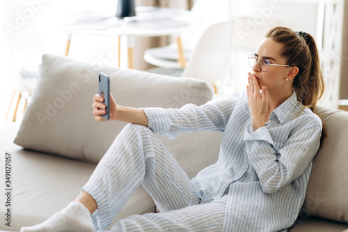 Phone video call. Attractive young woman communicates via video communication using a smartphone, sitting at home on the couch in her pajamas