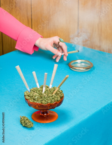 Hand in Pink Sleeve Holding Smoking Joint with Dish Full of Cannabis and Joints in Foreground on Blue Tablecloth (ID: 349046681)