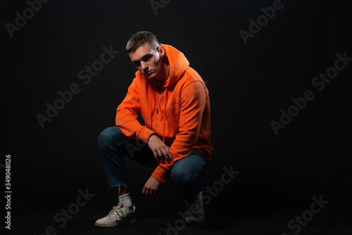 Male model sitting on the floor and posing in orange sports hoodie isolated over a black background.