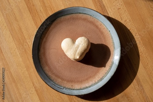 Heart shaped French macaron, cream colour, on a brown and blue clay plate. The small light and airy meringue cookie has a swirl in the crunchy exterior. The plate is sitting on an oak wooden table. 