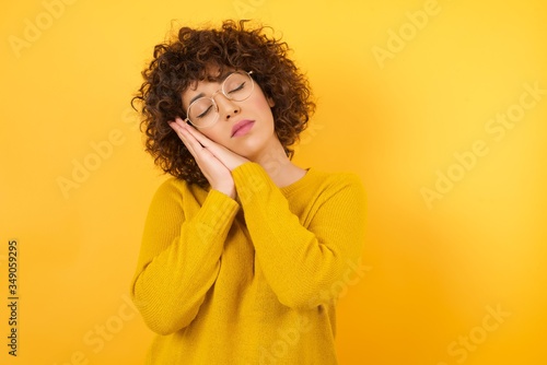 Young beautiful African American businesswoman with curly hair wearing yellow sweater  sleeping tired dreaming and posing with hands together while smiling with closed eyes. © Roquillo